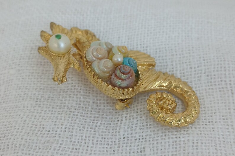 May include: A gold-tone seahorse brooch with a pearl and green stone eye. The seahorse is holding a cluster of iridescent seashells.