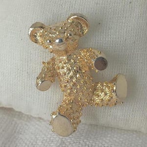 May include: A gold-coloured teddy bear brooch with a textured surface. The bear is depicted in a playful pose, with one leg raised. The brooch has silver accents on the nose and ears.
