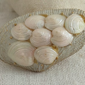 May include: A white and gold seashell brooch featuring a cluster of iridescent white seashells arranged in a heart shape.