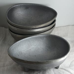 May include: A stack of five gray metal bowls with a textured surface. The bowls have a rounded, teardrop shape and sit on three small legs. The bowls have a silver rim.