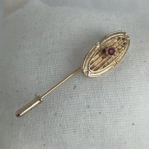 Peut inclure: Épingle de revers dorée avec une tête ovale ornée d'une pierre rouge centrale. L'épingle présente un motif texturé et une longue tige fine. Le bijou est probablement vintage ou antique.