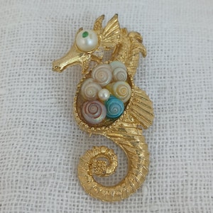 May include: Gold-tone seahorse brooch with a detailed design. The seahorse's body is filled with a cluster of colorful and iridescent seashells.