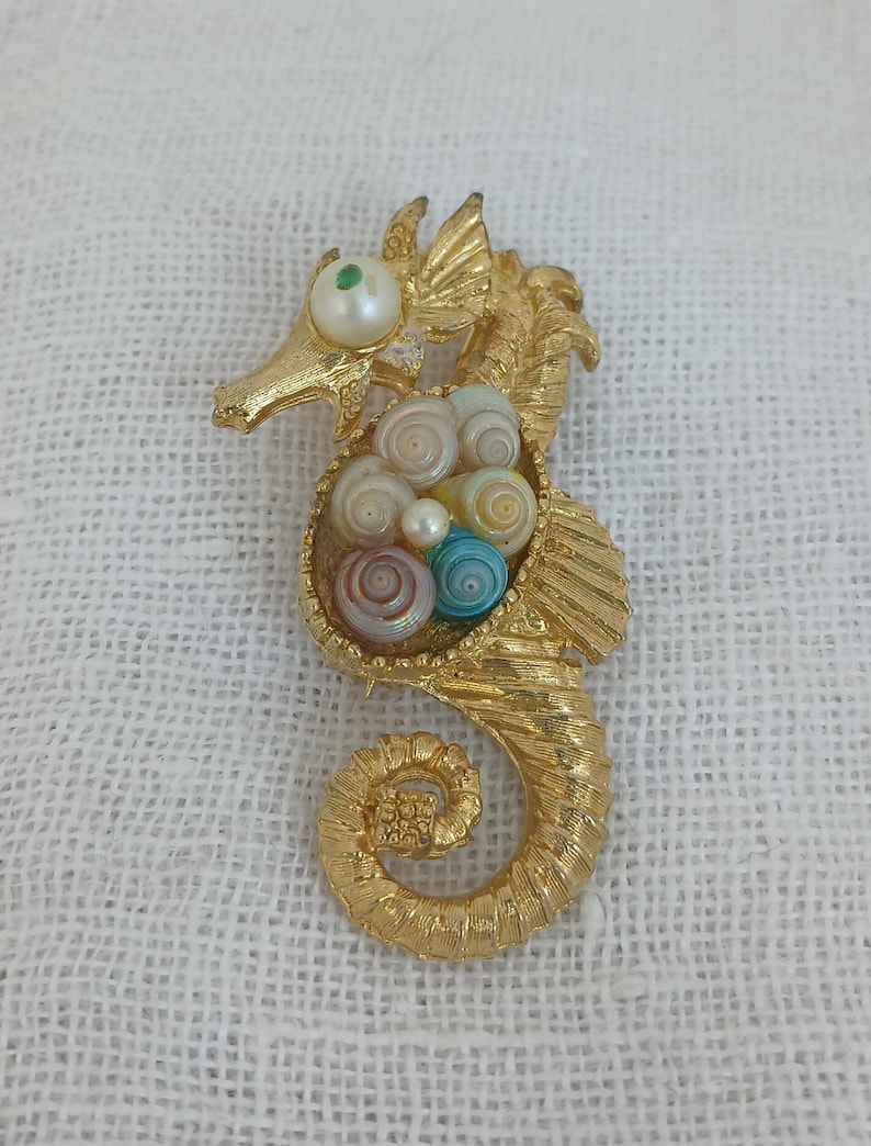 May include: Gold-tone seahorse brooch with a detailed design. The seahorse's body is filled with a cluster of colorful and iridescent seashells.