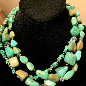 Puede incluir: Un collar de tres hilos con piedras de color turquesa de varios tamaños y formas. El collar tiene detalles de cristal transparente y se exhibe sobre un busto de terciopelo negro. Las piedras tienen tonos marrones y verdes.