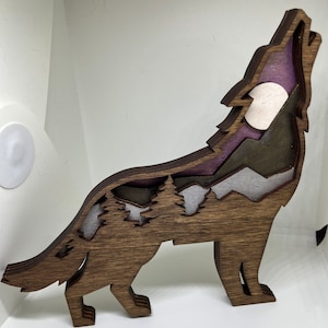May include: A wooden wolf silhouette with a layered mountain and forest scene inside. The wolf is howling at the moon, which is also cut out of the wood.