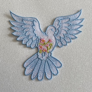 May include: Embroidered patch featuring a dove with outstretched wings. The dove is white with light blue outlining and wings. The dove's chest has a floral design with pink and yellow flowers. The patch is designed for sewing or ironing onto fabric.