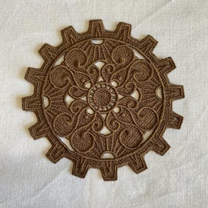 May include: A brown, decorative gear-shaped applique with intricate, swirling patterns. The applique is made of a textured material and features a central circular design. The gear has multiple teeth around its perimeter.