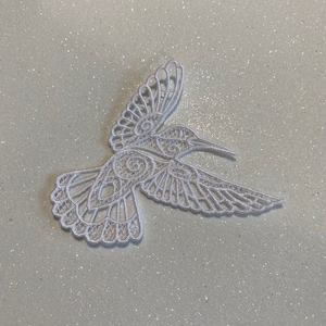 May include: A white embroidered hummingbird patch featuring intricate openwork lace details, swirling patterns within the wings, and a fanned tail design. The embroidery is crafted with fine white thread, creating a delicate, lightweight, and decorative bird motif.