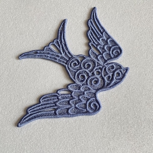 May include: A periwinkle blue embroidered swallow patch featuring intricate swirling patterns and detailed wing feathers. The bird is depicted in a flying position with its wings spread and tail feathers fanned out, showcasing a delicate lace-like texture.