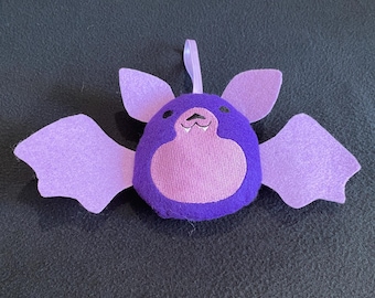 May include: A plush bat ornament featuring a deep purple body with a lighter lavender belly, ears, and wings. The bat has small black eyes, a stitched nose, and two white fangs. A lavender ribbon loop is attached to the top for hanging purposes.