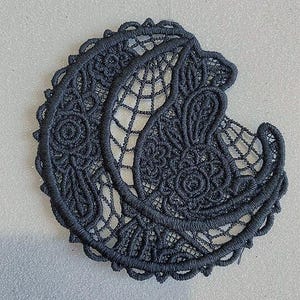 May include: A dark gray, crescent-shaped embroidered patch with intricate floral and geometric lace patterns. The patch features a scalloped edge and a delicate, open-weave design, suitable for embellishing clothing or accessories.