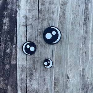 May include: Three black and white circular embellishments with a crescent moon and two white dots design. The embellishments are in varying sizes and are arranged on a weathered wooden surface.
