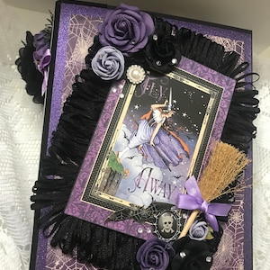 May include: Purple and black scrapbook with a witch flying on a broomstick, surrounded by black and purple flowers and a skull and crossbones charm. The scrapbook has a black fringe and a purple ribbon.