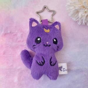 Può includere: Portachiavi a forma di gatto di peluche viola con un design a mezzaluna e stella. Il portachiavi ha un anello in metallo argentato.
