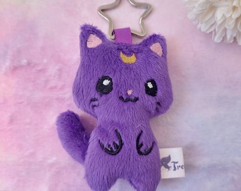 Puede incluir: Llavero de gato de peluche morado con un diseño de luna creciente y estrella. El llavero tiene un anillo de metal plateado.
