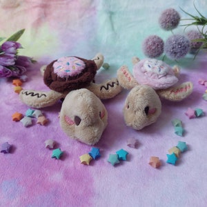 Può includere: Due giocattoli di peluche a forma di tartaruga con gusci marroni e rosa. I gusci sono decorati con un design a ciambella. Le tartarughe sono sedute su una superficie pelosa viola e blu con stelle di origami colorate.
