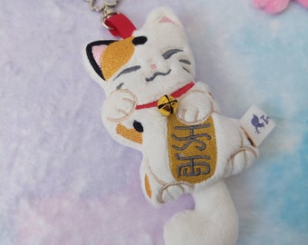 May include: White plush Maneki-Neko, or lucky cat, keychain with a gold bell and a red collar. The cat is holding a gold sign with Japanese characters.