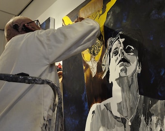 Op de afbeelding: Een kunstenaar in een witte jas schildert op een groot canvas. Het kunstwerk toont een grijswaardenportret van een persoon die omhoog kijkt, met een donkerblauwe en gele achtergrond. De kunstenaar gebruikt een penseel.