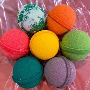 May include: A collection of seven colorful bath bombs in various shades, including green, orange, red, yellow, teal, and purple. Each bath bomb is spherical, with some featuring a textured band around the middle. The bath bombs are arranged on a pink, crinkled surface.