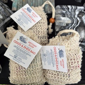 May include: Several beige mesh bags labeled "Odds & Ends Bag" filled with colorful soap pieces. Each bag has a drawstring closure with a wooden bead. The bags are displayed on a dark surface, with product tags attached.