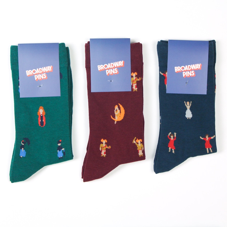 May include: Three pairs of socks with a Broadway Pins logo. The socks are green, burgundy, and navy blue. Each pair features a different Broadway show theme with pixelated images of characters and scenes.