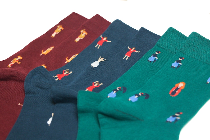 May include: Three pairs of socks with different patterns. One pair is burgundy with a yellow dog pattern, one pair is dark blue with a white dancing woman pattern, and one pair is green with a blue and red woman pattern.