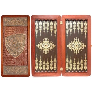 Könnte beinhalten: Ein Backgammon-Spiel aus Holz mit braunem, lederartigem Äußeren und aufwendigen goldfarbenen Designs. Das Spielbrett ist geschlossen und zeigt einen dekorativen Schild mit einem Löwenmotiv.