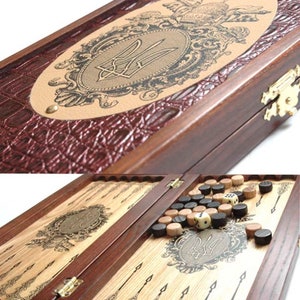 Könnte beinhalten: Ein Backgammon-Set aus Holz mit braunem, lederartigem Äußeren. Die Oberseite der Box zeigt ein goldenes Emblem mit einem Dreizack-Design. Die Innenseite der Box ist geöffnet und zeigt das Spielbrett und schwarze und braune Spielsteine.
