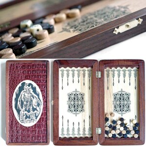 Könnte beinhalten: Ein Backgammon-Set aus Holz mit roter Lederausstattung und einem Wikingerkrieger-Design. Das Set ist geöffnet und zeigt die Spielfläche mit schwarzen und weißen Spielsteinen.