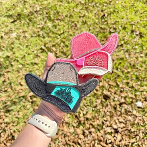 May include: Two cowboy hat-shaped resin coasters. One is black with a turquoise band and a floral design, the other is pink with a silver glitter brim. Both have a glittery outline and are held against a background of green grass and fallen leaves.