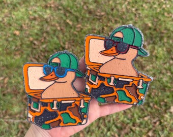 May include: Two duck-shaped accessories, each featuring a duck wearing sunglasses and a green cap, inside a camouflage-patterned box. The ducks have orange beaks and are set against a background of green grass.