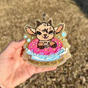 May include: A whimsical goat-themed charm featuring a cartoon goat floating on a pink donut with sprinkles. The charm has a gold glittery border and the text "WHATEVER FLOATS YOUR GOAT".