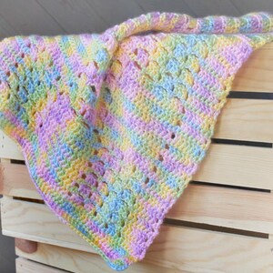 Puede incluir: Una manta de bebé de crochet multicolor en tonos de rosa, amarillo, verde y azul. La manta está colocada sobre una caja de madera.