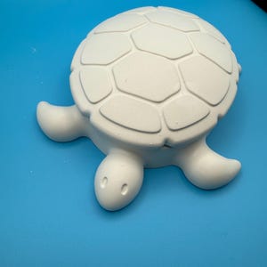 May include: A white, unpainted ceramic turtle figurine featuring a smooth, rounded shell with a raised, segmented geometric pattern. The turtle has a simple head with two small, oval-shaped indentations for eyes and four extended flipper-like limbs.