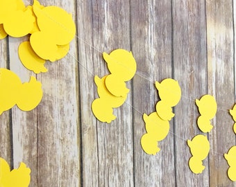 May include: A yellow paper garland with duck shapes strung together on a white string. The garland is hanging against a wood background.