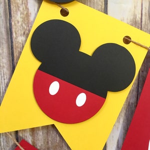 May include: Yellow pennant banner with a black and red Mickey Mouse head silhouette. The head is outlined in black and has two white circles for eyes.