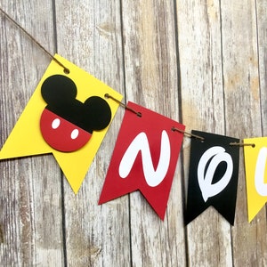 K&ouml;nnte beinhalten: Ein gelbes, rotes und schwarzes Banner mit dem Wort "NOEL" in wei&szlig;en Buchstaben. Das Banner hat auf der linken Seite einen Mickey-Maus-Kopf.
