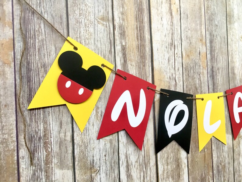 K&ouml;nnte beinhalten: Ein gelbes, rotes und schwarzes Banner mit dem Wort "NOEL" in wei&szlig;en Buchstaben. Das Banner hat auf der linken Seite einen Mickey-Maus-Kopf.