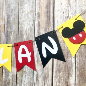 K&ouml;nnte beinhalten: Ein gelbes, rotes, schwarzes und wei&szlig;es Banner mit dem Wort "PLAN" in wei&szlig;en Buchstaben. Das Banner hat einen Mickey Mouse Kopf auf dem gelben Teil ausgeschnitten.