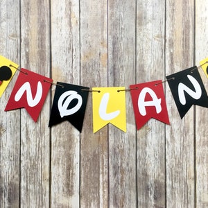 K&ouml;nnte beinhalten: Ein gelbes, rotes und schwarzes Banner mit dem Namen "NOLAN" in wei&szlig;en Buchstaben. Jeder Buchstabe befindet sich auf einem separaten Wimpel. Das Banner ist mit Mickey-Maus-K&ouml;pfen verziert.