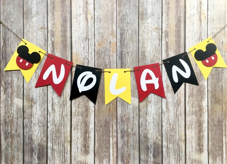 K&ouml;nnte beinhalten: Ein gelbes, rotes und schwarzes Banner mit dem Namen "NOLAN" in wei&szlig;en Buchstaben. Jeder Buchstabe befindet sich auf einem separaten Wimpel. Das Banner ist mit Mickey-Maus-K&ouml;pfen verziert.