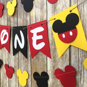Puede incluir: Una guirnalda decorativa con banderines de papel en rojo, amarillo y negro. Tres banderines centrales muestran las letras blancas O, N y E. Otros banderines con forma de orejas de rat&oacute;n se intercalan a lo largo de la cuerda en un patr&oacute;n.