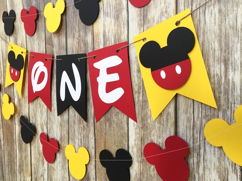 Puede incluir: Una guirnalda decorativa con banderines de papel en rojo, amarillo y negro. Tres banderines centrales muestran las letras blancas O, N y E. Otros banderines con forma de orejas de rat&oacute;n se intercalan a lo largo de la cuerda en un patr&oacute;n.