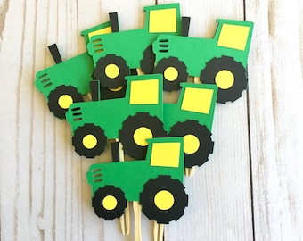 May include: Green and yellow tractor cupcake toppers. The toppers are made of cardstock and have a black outline. They are attached to wooden sticks.