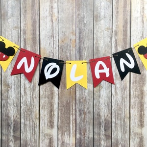 K&ouml;nnte beinhalten: Ein gelbes, rotes und schwarzes Banner mit dem Namen "NOLAN" in wei&szlig;en Buchstaben. Jeder Buchstabe befindet sich auf einem separaten Wimpel mit einem Mickey-Maus-Kopf an jedem Ende des Banners.