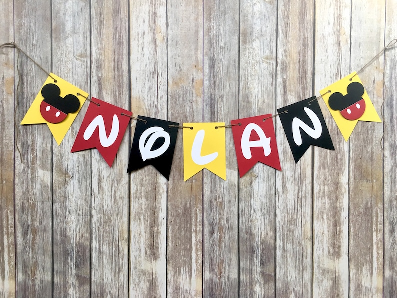 K&ouml;nnte beinhalten: Ein gelbes, rotes und schwarzes Banner mit dem Namen "NOLAN" in wei&szlig;en Buchstaben. Jeder Buchstabe befindet sich auf einem separaten Wimpel mit einem Mickey-Maus-Kopf an jedem Ende des Banners.