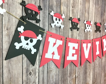Peut inclure: Une bannière de pirate rouge et blanche avec le nom "KEVIN" en lettres blanches sur des drapeaux rouges et des crânes et des os croisés noirs et blancs sur des drapeaux noirs.