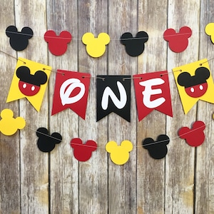 Puede incluir: Una guirnalda decorativa con la palabra ONE en letras blancas sobre banderines rojos y negros, flanqueada por banderines amarillos con cabezas de Mickey Mouse. La guirnalda incluye recortes adicionales de cabezas de Mickey en rojo, amarillo y negro.