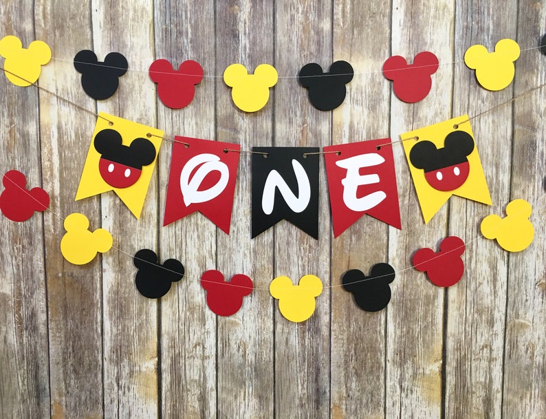 Puede incluir: Una guirnalda decorativa con la palabra ONE en letras blancas sobre banderines rojos y negros, flanqueada por banderines amarillos con cabezas de Mickey Mouse. La guirnalda incluye recortes adicionales de cabezas de Mickey en rojo, amarillo y negro.