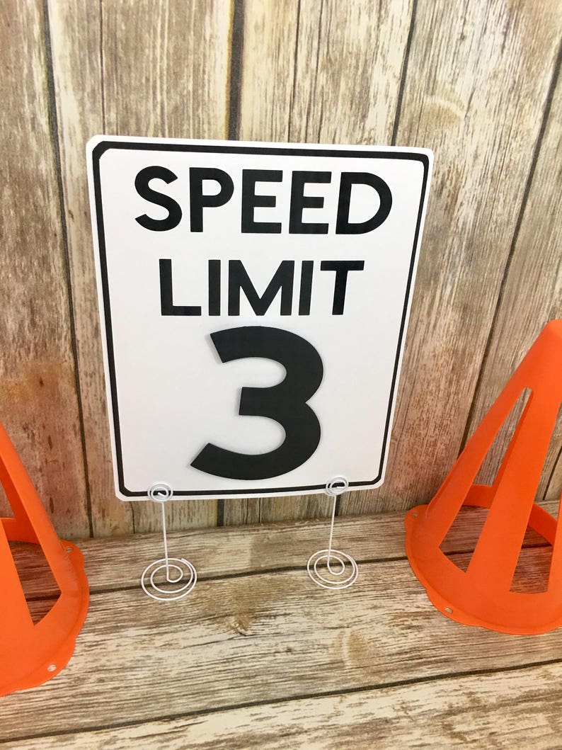 May include: White speed limit sign with black text that reads "SPEED LIMIT 3". The sign is on a white stand with a spiral base. Two orange traffic cones are on either side of the sign.
